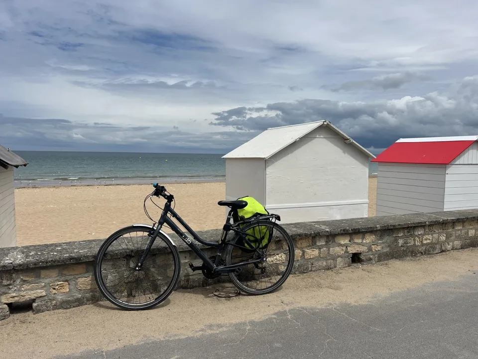 Vélo avec sac jaune fluo adossé à un muret devant des cabines de plage blanches et rouges, avec la mer et un ciel nuageux en arrière-plan